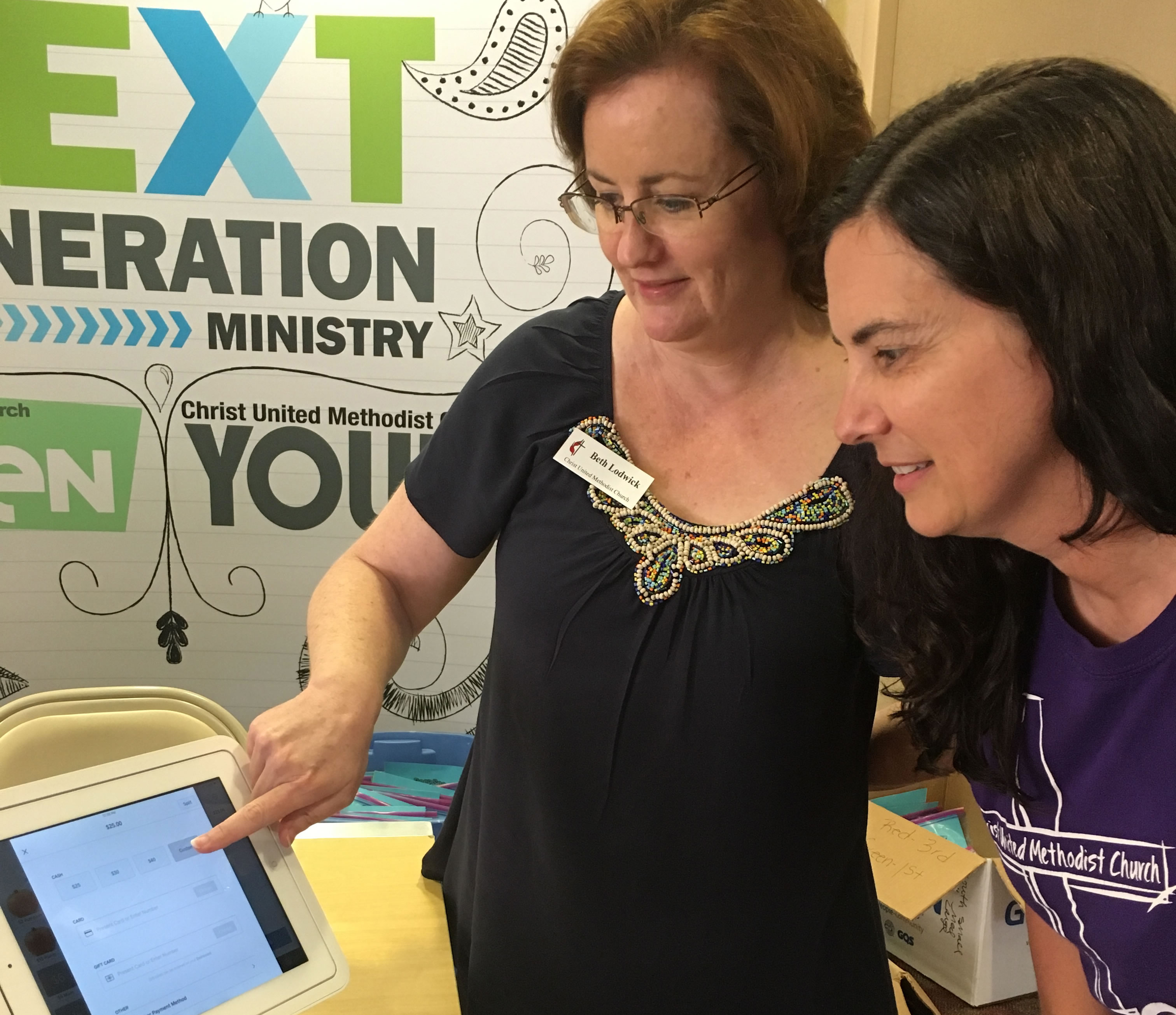Beth Lodwick (left), finance and youth assistant at Christ United Methodist Church in Mobile, Ala., demonstrates use of a Square tablet to church member Angela Tait. The device lets buyers use credit and debit cards at church fundraisers. The church also promotes online giving of church offerings. Photo courtesy of Christ United Methodist Church, Mobile, Ala.