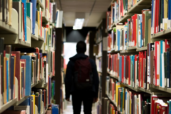 Student walking through library stacks. Photo by Banter Snaps, Unsplash.com.