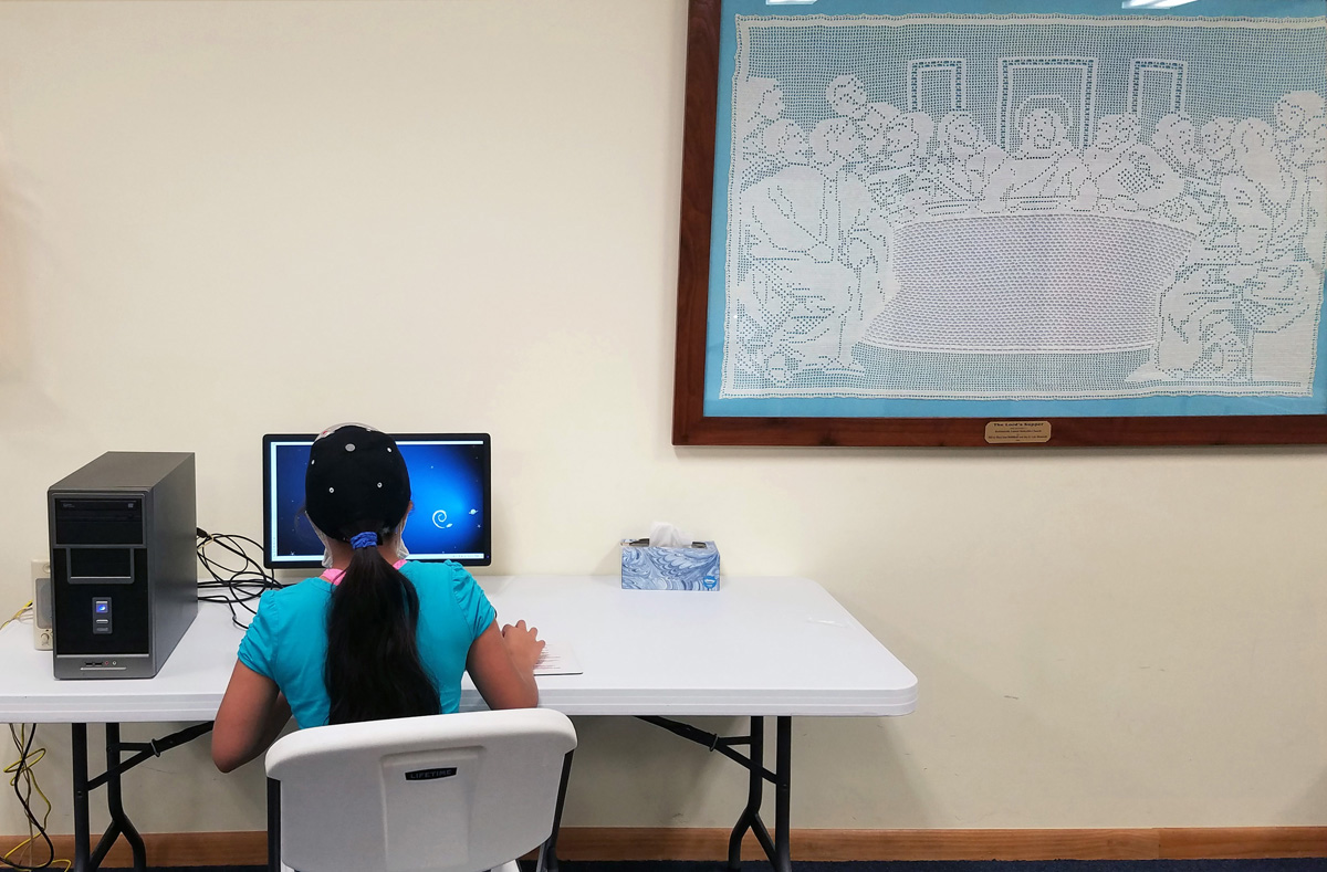 Sarai Moreno, a rising seventh grader, does homework on a computer at Robbinsville United Methodist Church in North Carolina. The church has worked to provide access to remote learning in a rural county where more than half of households have no internet. Women in the church created the needlepoint Last Supper at right. Photo by the Rev. Eric Reece.
