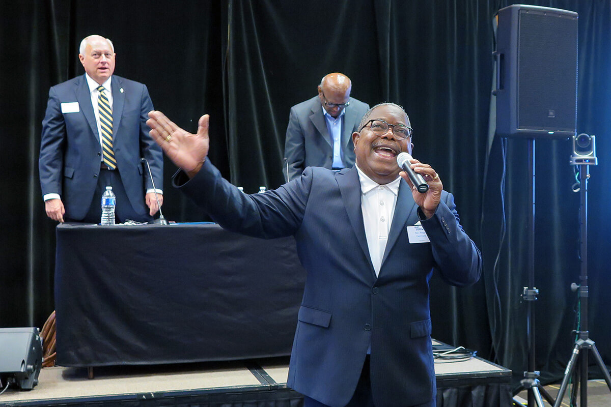 The Rev. Reggie Clemmons, vice president of the General Council on Finance and Administration board, leads morning devotion Aug. 7 at the board’s organizing meeting in Franklin, Tenn. He is singing “How Great Is Our God.” Behind him are Bishop David Graves (left), GCFA board chair, and the Rev. Moses Kumar, the top executive of the denomination’s finance agency. Photo by Heather Hahn, UM News.
