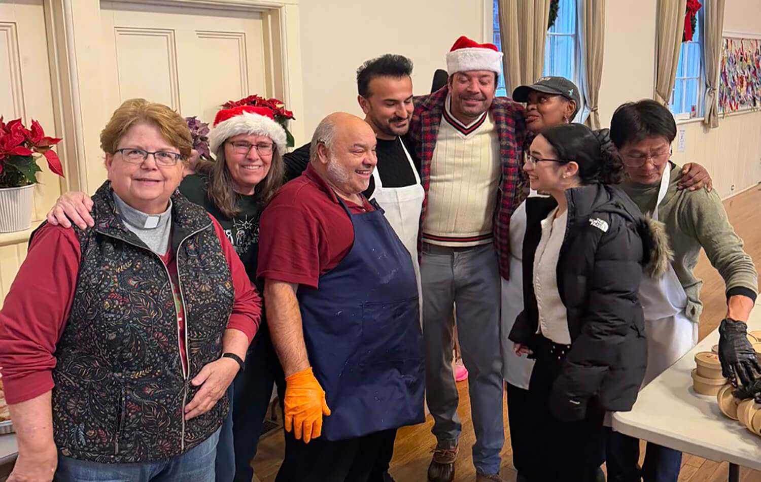 La Revda. Joanne Utley (izquierda) junto  un grupo de voluntarios/as rodeando al comediante Jimmy Fallon, quien se presentó en la IMU de Hamptons junto a su familia, para servir alimentos el día de Navidad. Foto cortesía de la IMU de Hamptons.