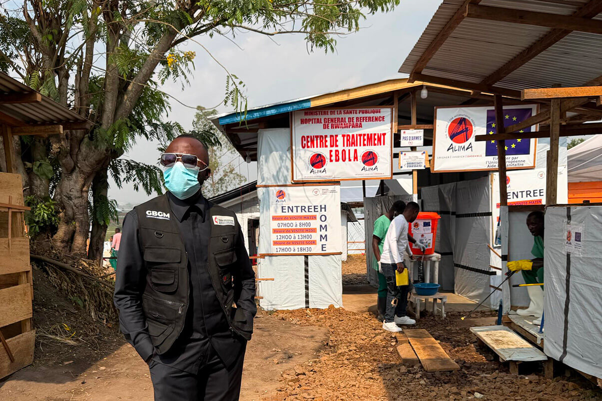 Dr. Damas Lushima, wearing a mask and a Global Ministries vest, stands in front of the Ebola treatment center at the Rwampara General Referral Hospital in Bunia. As health department coordinator for the East Congo Episcopal Area, he oversees The United Methodist Church’s integrated response to the 17th Ebola outbreak affecting Ituri Province. Photo courtesy of the Eastern Congo Health Council.
